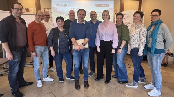 Gruppe von Personen steht in einem Seminarraum vor einer Leinwand, auf der eine Präsentation angezeigt wird.