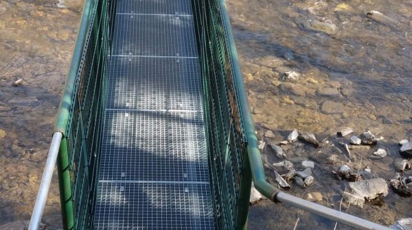 Schmale Metall-Hängebrücke mit grünen seitlichen Geländern, die über einen flachen, steinig fließenden Bach führt.