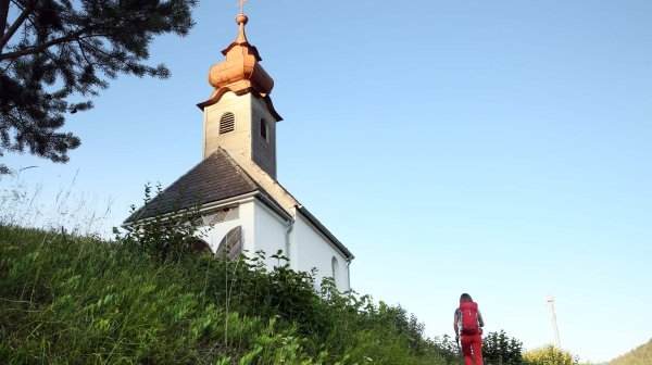 Kleine Kapelle mit hellem Mauerwerk und einem zwiebelförmigen Turmaufsatz auf einem Hügel, umgeben von grüner Vegetation. Auf einem schmalen Weg führt eine wandernde Person mit rotem Outfit und Rucksack den Hügel hinauf zur Kapelle. Der Himmel ist klar und blau.
