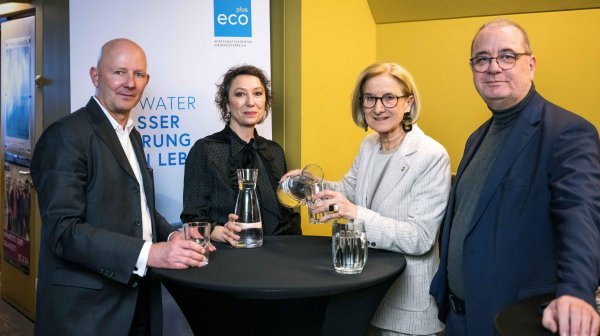 Eine Gruppe von vier Personen steht an einem hohen Stehtisch in einem Innenraum. Auf dem Tisch befinden sich mehrere Gläser und eine Glaskaraffe mit Wasser.