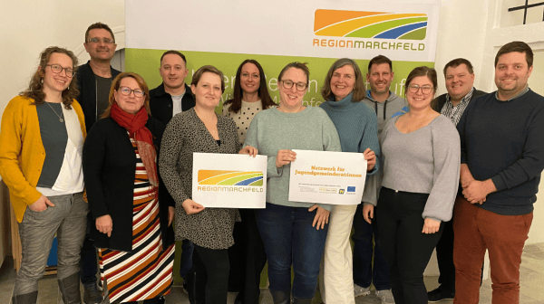 Eine Gruppe von dreizehn Personen steht gemeinsam vor einem Roll‑Up der „Region Marchfeld“. Zwei Frauen in der Mitte halten Schilder hoch – eines mit dem Logo der Region Marchfeld, das andere mit dem Text „Netzwerk für Jugendgemeinderät:innen“. Alle Personen lächeln in die Kamera. 