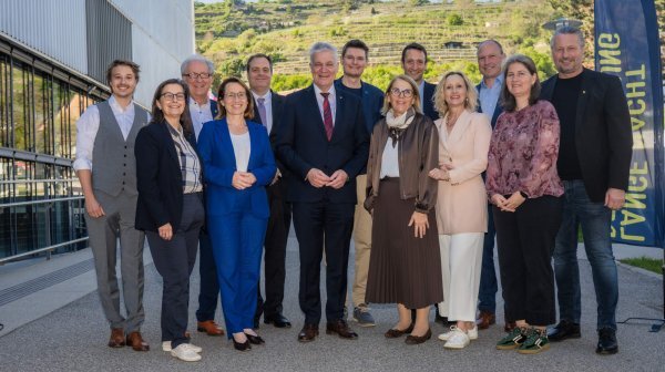 Eine Gruppe von vierzehn erwachsenen Personen steht in zwei Reihen im Freien vor einem modernen Gebäude.