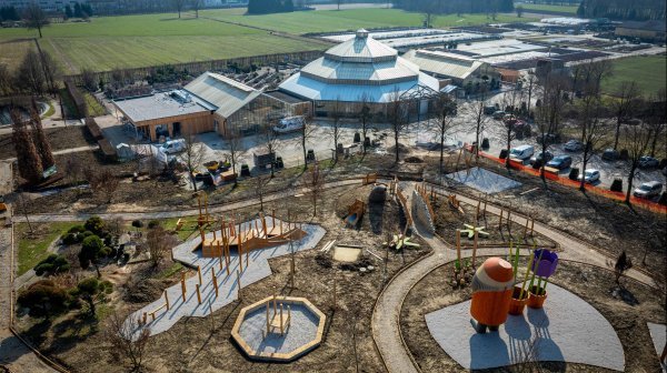 Luftaufnahme einer neu gestalteten Park- und Spiellandschaft mit geschwungenen Wegen, Holzspiel- und Kletterelementen sowie bunten Skulpturen, im Hintergrund moderne Gewächshausgebäude und weitläufige Grünflächen in einer offenen Landschaft.