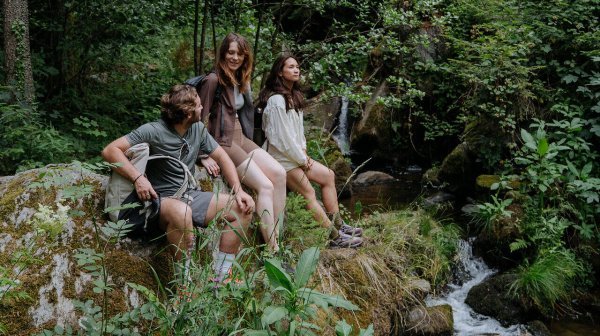 Drei junge Erwachsene sitzen bei einer Wanderpause an einem kleinen Bach in einem dichten, grünen Wald. 