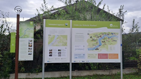 Informationstafeln mit Karten und Erläuterungen zu einer Region stehen im Freien vor einem Gebäude, umgeben von Grünflächen und Bäumen unter bewölktem Himmel.