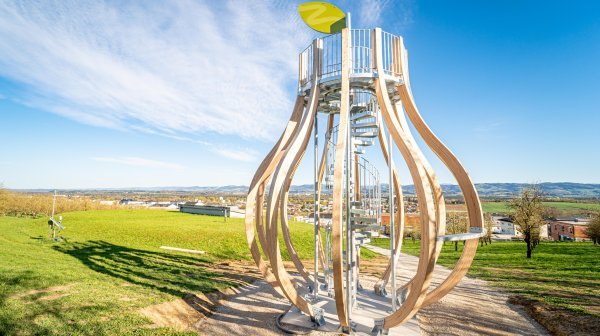 Hölzerner Aussichtsturm mit spiralförmiger Treppe und blattförmigem Dach inmitten einer Obstbaumallee, davor grünes Gras und ein Weg, darüber blauer Himmel.