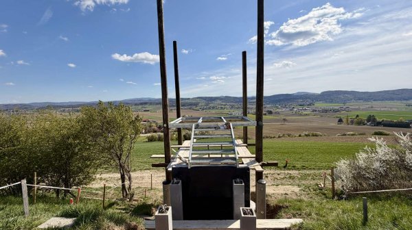 Holz- und Stahlkonstruktion eines im Bau befindlichen Aussichtspunktes auf einem Hügel, mit freiem Blick über Felder, Dörfer und sanfte Hügellandschaft unter blauem Himmel.