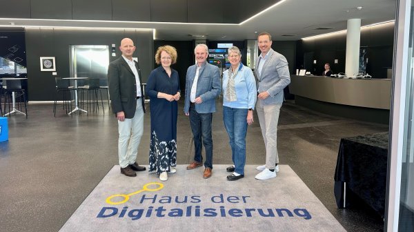 Fünf erwachsene Personen stehen nebeneinander in der modernen Eingangshalle. Sie stehen auf einer Bodenmatte mit der Aufschrift „Haus der Digitalisierung“. 