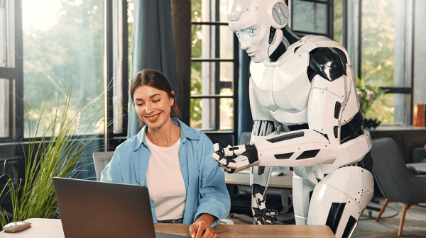 Dame sitzt am Computer und ein Roboter schaut ihr über die Schulter und erklärt ihr etwas