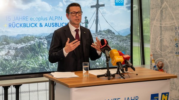 Ein Mann in Anzug und Brille bei einer Pressekonferenz vor einem Screen mit einem Bergfoto