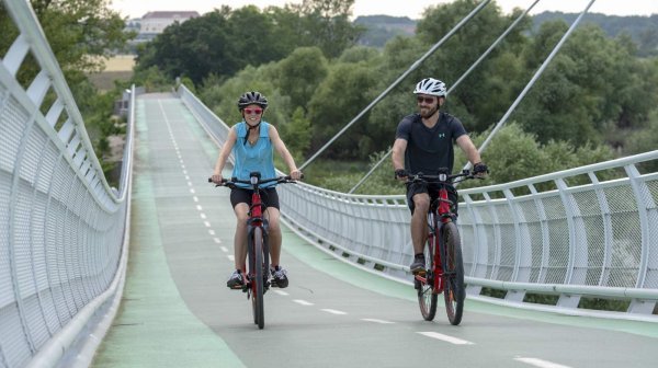 Zwei Personen fahren mit Fahrrädern über eine moderne, leicht geschwungene Fuß- und Radbrücke mit Geländern und Seilabspannungen; im Hintergrund sind Bäume und eine grüne Flusslandschaft zu sehen.