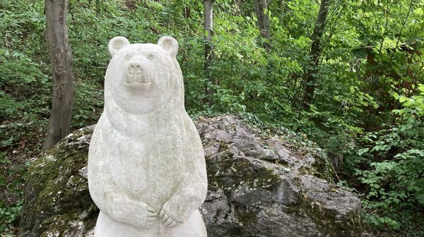 Steinerne Skulptur eines aufrecht stehenden Braunbären auf einem Felsblock in einem bewaldeten Gebiet.