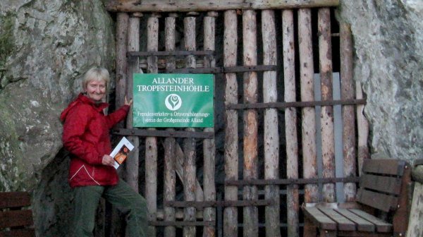 Eingang einer Höhle in einer Felswand, verschlossen durch ein einfaches Gitter aus dicken Holzstangen.