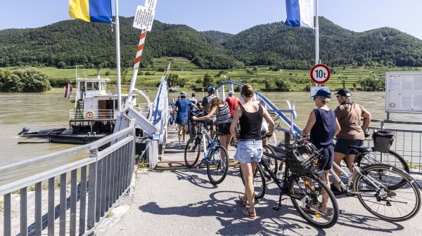 Mehrere Personen schieben Fahrräder auf eine kleine Fähre über die Donau. Im Vordergrund führt ein Steg mit Geländern zur Fähre.