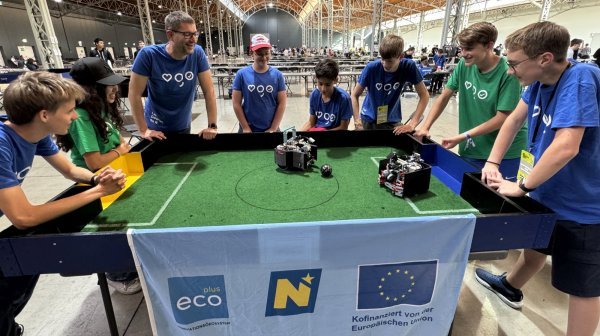 Jugendliche bei einem Roboterfußball-Bewerb stehen rund um ein Spielfeld in einer großen Veranstaltungshalle und beobachten zwei kleine Roboter, die mit einem Ball spielen.