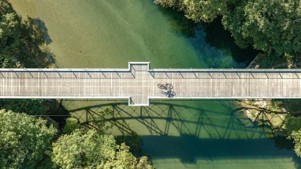 Luftaufnahme einer geraden Holzbrücke, die über einen klaren, grünlich schimmernden Fluss führt. In der Mitte der Brücke stehen zwei Fahrräder nebeneinander. 
