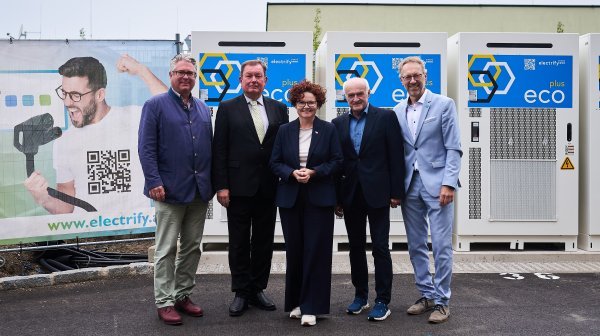 Fünf Personen stehen vor Schwarmspeichergeräten mit ecoplus und Electrify Logos