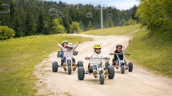 Kinder fahren mit Mountaincarts auf einem Schotterweg durch eine grüne Berglandschaft unter einem Sessellift.