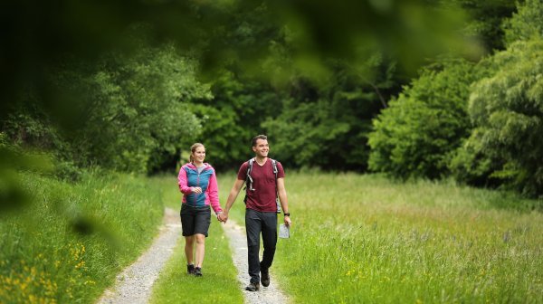Zwei Personen mit Wanderausrüstung gehen auf einem schmalen Schotterweg durch eine grüne Wiesenlandschaft. Eine Person trägt einen Rucksack und hält eine Wanderkarte in der Hand, die andere trägt eine Outdoorjacke. Im Hintergrund befindet sich dichter Baumbestand.