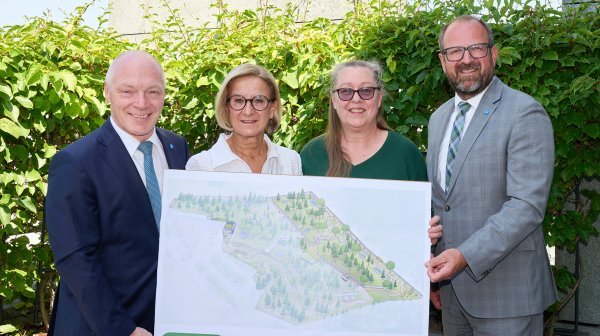 Vier erwachsene Personen stehen im Freien vor einer dichten grünen Hecke und halten gemeinsam eine große Präsentationstafel.