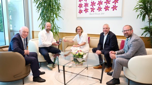 Fünf Personen sitzen in einer modernen Besprechungsecke um einen runden Glastisch. In der Mitte des Bildes wird ein mobiles Messgerät beziehungsweise technisches Handgerät übergeben.