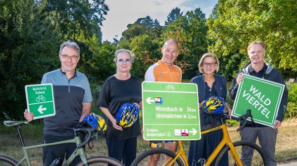 Fünf Personen stehen im Freien vor Bäumen und halten Verkehrsschilder zum Radwegenetz. Zwei Fahrräder sind im Vordergrund sichtbar.