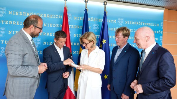 Fünf Personen stehen vor einer Fotowand, in der Mitte hält eine Person ein Dokument, das gemeinsam betrachtet wird.