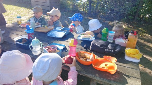Mehrere Kinder sitzen an einem Picknicktisch im Freien und essen gemeinsam. Auf dem Tisch stehen Teller, Getränke und verschiedene Speisen.