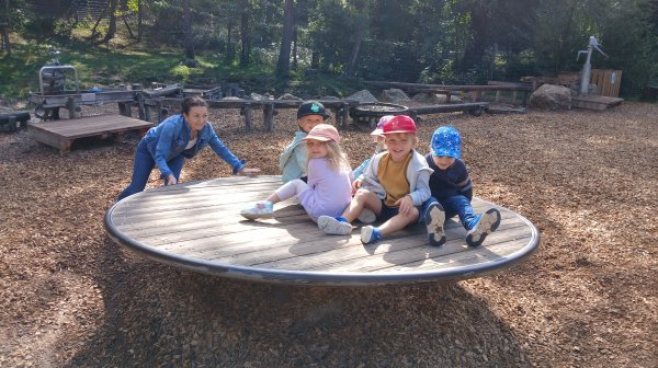 Mehrere Kinder spielen gemeinsam auf einem großen, runden Spielgerät auf einem Spielplatz im Freien. Der Boden ist mit Holzschnitzeln bedeckt, im Hintergrund sind Bäume und weitere Spielgeräte zu sehen.