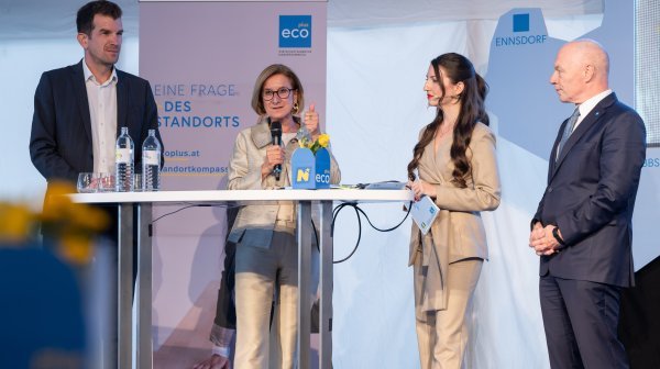 Drei Personen befinden sich an einem Stehtisch mit Mikrofonen und technischen Geräten, während eine weitere Person rechts daneben steht.