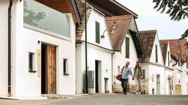 Historische Kellergasse mit weiß getünchten Häusern und ziegelgedeckten Dächern. Im Vordergrund ist ein modern saniertes Gebäude mit Holzfassade und Glasbalkon zu sehen. Zwei Personen gehen die leicht ansteigende Straße entlang.