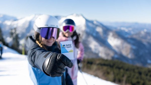 Zwei Wintersportlerinnen stehen auf einer verschneiten Skipiste vor einer Bergkulisse. Im Vordergrund zeigt eine Person mit ausgestrecktem Arm den Bergerlebnispass in die Kamera
