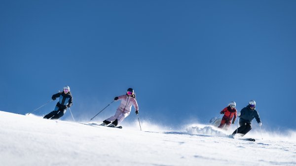 Vier Personen fahren auf Skiern eine breite, verschneite Piste einen Hang hinunter. Hinter den Personen wirbelt Schnee auf, darüber spannt sich ein klarer, wolkenloser Himmel