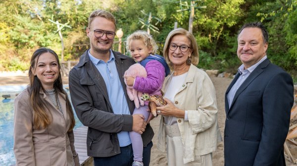 Fünf Personen stehen im Freien vor einer naturnah gestalteten Anlage mit Sandbereich und Wasserbecken. Eine Person hält ein kleines Kind auf dem Arm, das ein rosafarbenes Stofftier trägt.
