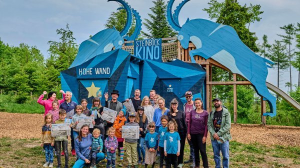 Eine Gruppe aus Erwachsenen und Kindern steht auf einer Wiese vor einer großen Holzwand mit dem Schriftzug „HOHE WAND“. Mehrere Personen halten Landkarten in die Kamera.
