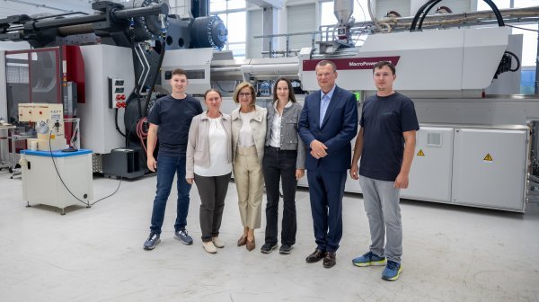 Sechs Personen stehen vor einer großen Maschine in einer Fabrikhalle, zwei Männer links, drei Frauen und ein Mann rechts.