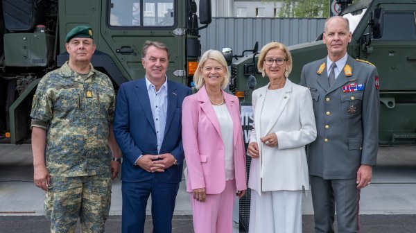 Fünf Personen stehen nebeneinander vor Militärfahrzeugen im Außenbereich. Zwei Personen tragen Uniformen, drei sind in ziviler Kleidung gekleidet. Die Gruppe blickt in die Kamera und posiert für ein gemeinsames Foto.