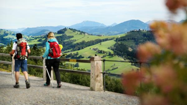 Zwei Personen wandern entlang eines Pflasterweges und im Hintergrund ist eine hügelige Landschaft. 