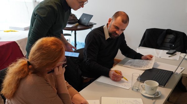 Drei Personen beugen sich über Zettel auf Tisch